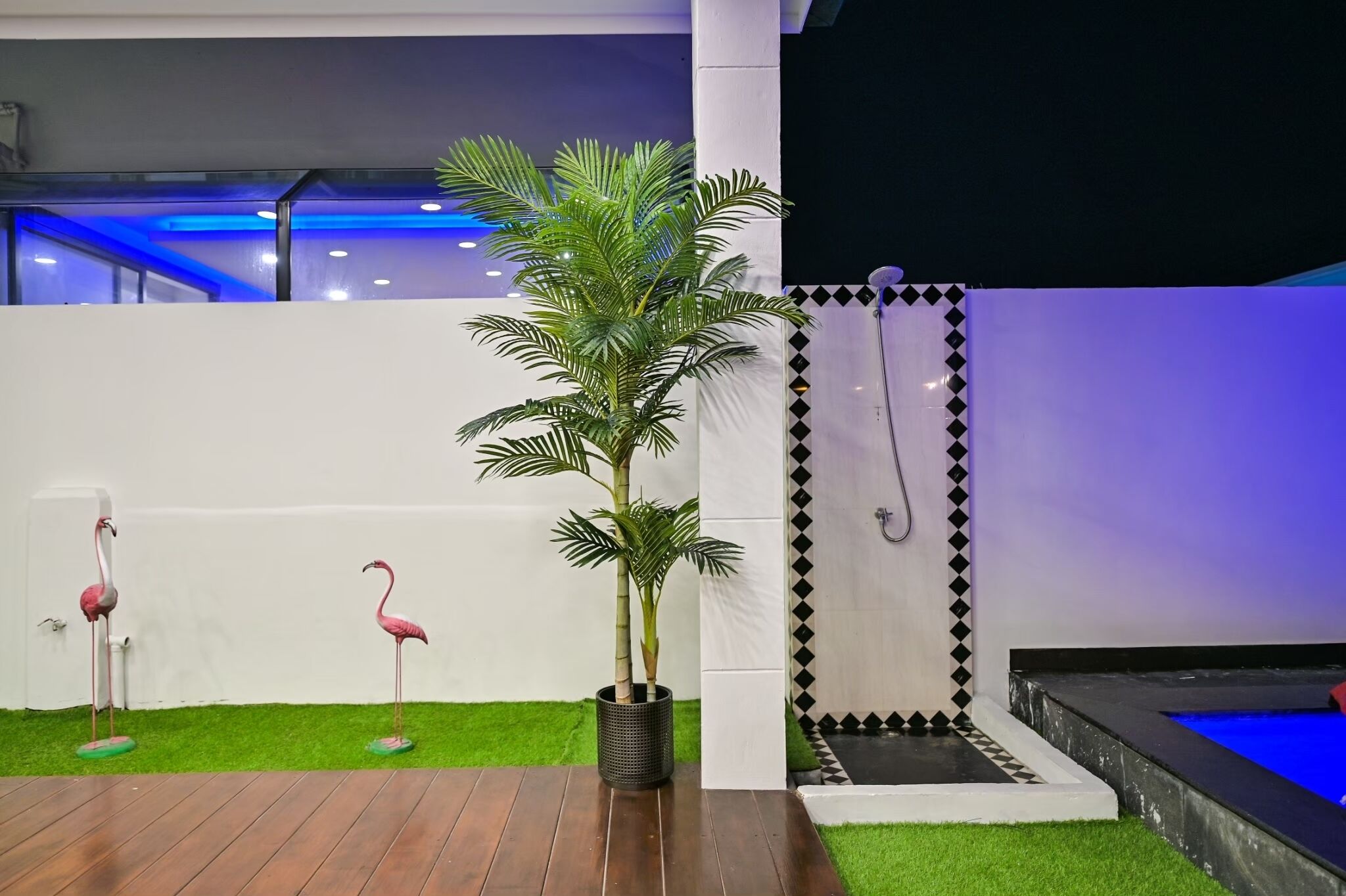 Un espace extérieur moderne avec une terrasse en bois, des flamants roses artificiels, un palmier, un mur carrelé en noir et blanc et une piscine bleue.