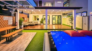 A modern indoor pool area with a glass ceiling, wooden benches, and a small seating area. - Exquisite Pool Villa A  (Pattaya)
