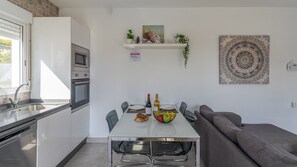 Uma cozinha moderna com uma mesa de jantar, cadeiras, um sofá cinzento e uma prateleira fixada na parede com objetos decorativos.
