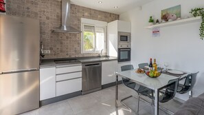 Uma cozinha moderna com geladeira, lava-louças e forno de aço inoxidável, uma área de refeições com mesa e cadeiras de vidro, e uma janela que oferece iluminação natural.