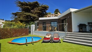 A modern house with a swimming pool, two lounge chairs, and a patio area with a table and chairs.