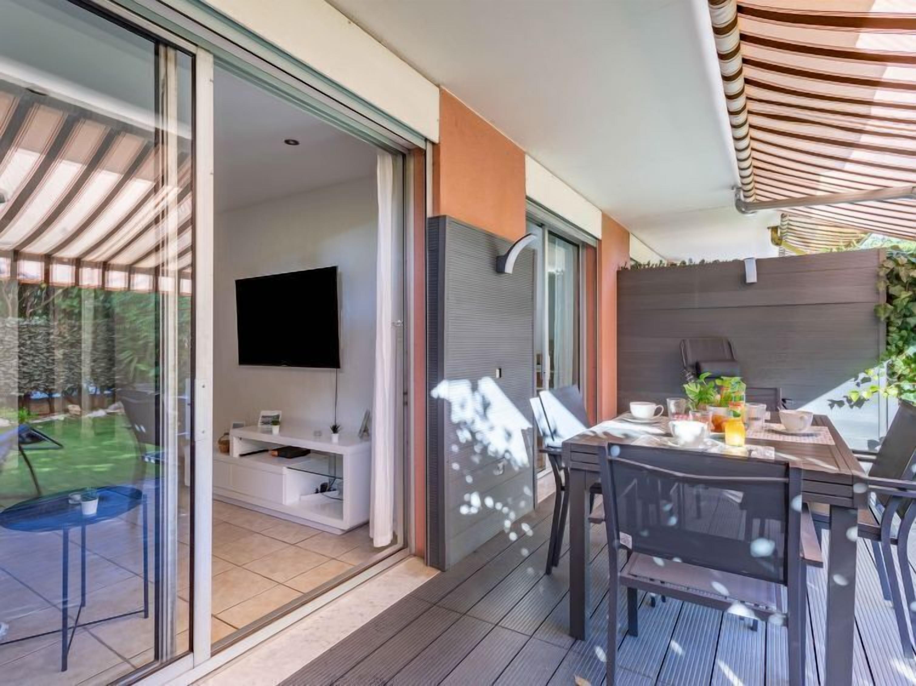 Uma área moderna para refeições ao ar livre, com mesa e cadeiras, uma televisão na parede e uma porta de vidro deslizante para um jardim.