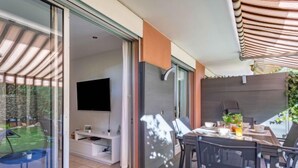 Uma área moderna para refeições ao ar livre, com mesa e cadeiras, uma televisão na parede e uma porta de vidro deslizante para um jardim.