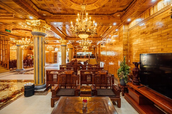 A spacious lobby with ornate wooden columns, a large chandelier, and a television on the right.