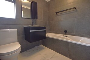 Features a sleek vanity, large mirror, white toilet, and spacious bathtub with a black faucet.