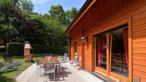Ein Holzgebäude mit einer Terrassenfläche, komplett mit Tisch, Stühlen, einem Backofen aus Ziegeln und einer Glastür zu einem Sitzbereich im Freien.