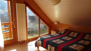 Ein Schlafzimmer mit Holzboden, einem Bett mit einem gemusterten Bettbezug, einem Fenster mit Blick ins Freie und einem kleinen Nachttisch mit Lampe.