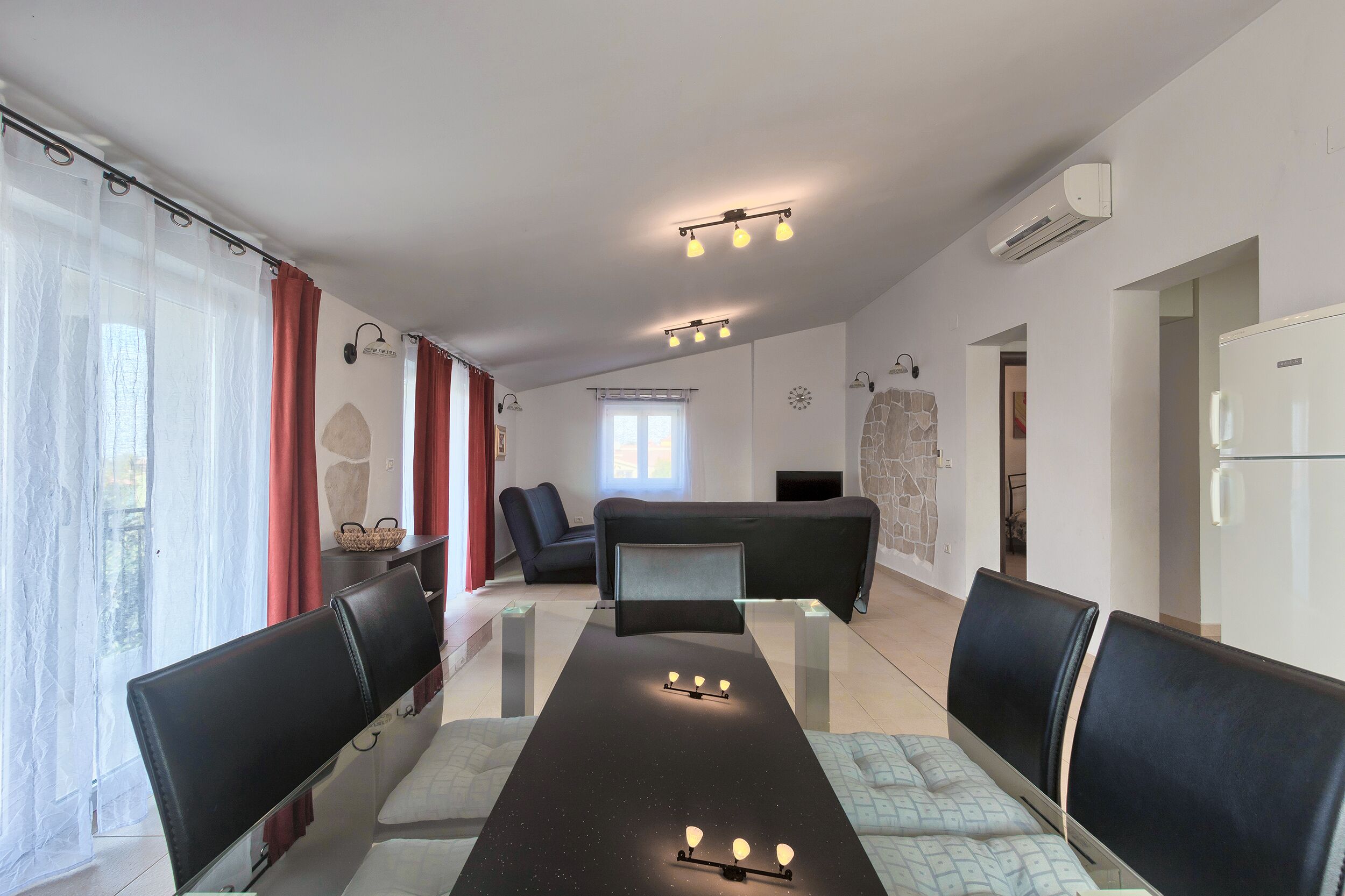 A modern room with a long table, black chairs, and a sofa. There are ceiling lights and a wall-mounted air conditioner.