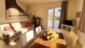 Un salon avec un canapé, une table à manger ornée de fruits, un téléviseur et un balcon offrant une vue agréable.