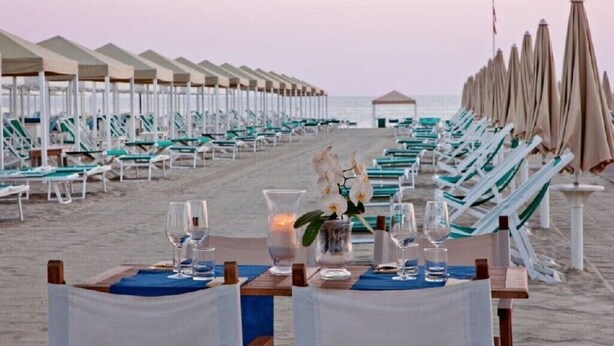 Un espace de restauration aménagé au bord de la plage, avec des rangées de chaises de plage et de parasols, une table dressée avec des verres et une bougie, et une vue sur l’océan en arrière-plan.