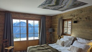 Une chambre avec un lit, une chaise, une lampe et une vue sur les montagnes par la fenêtre.