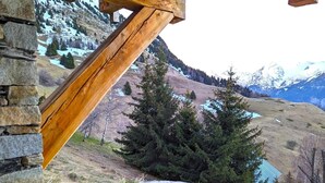 Un mur de pierres avec une structure en poutres de bois, un sentier pavé de pierres et une vue sur un paysage montagneux aux sommets enneigés et un petit lac.