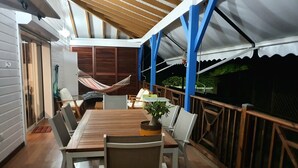 A covered outdoor dining area with a wooden table and chairs, a hammock, and a view of greenery.