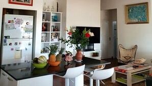 A modern kitchen with a central island, white bar stools, a dining area with a table and chairs, a television, and a bookshelf.