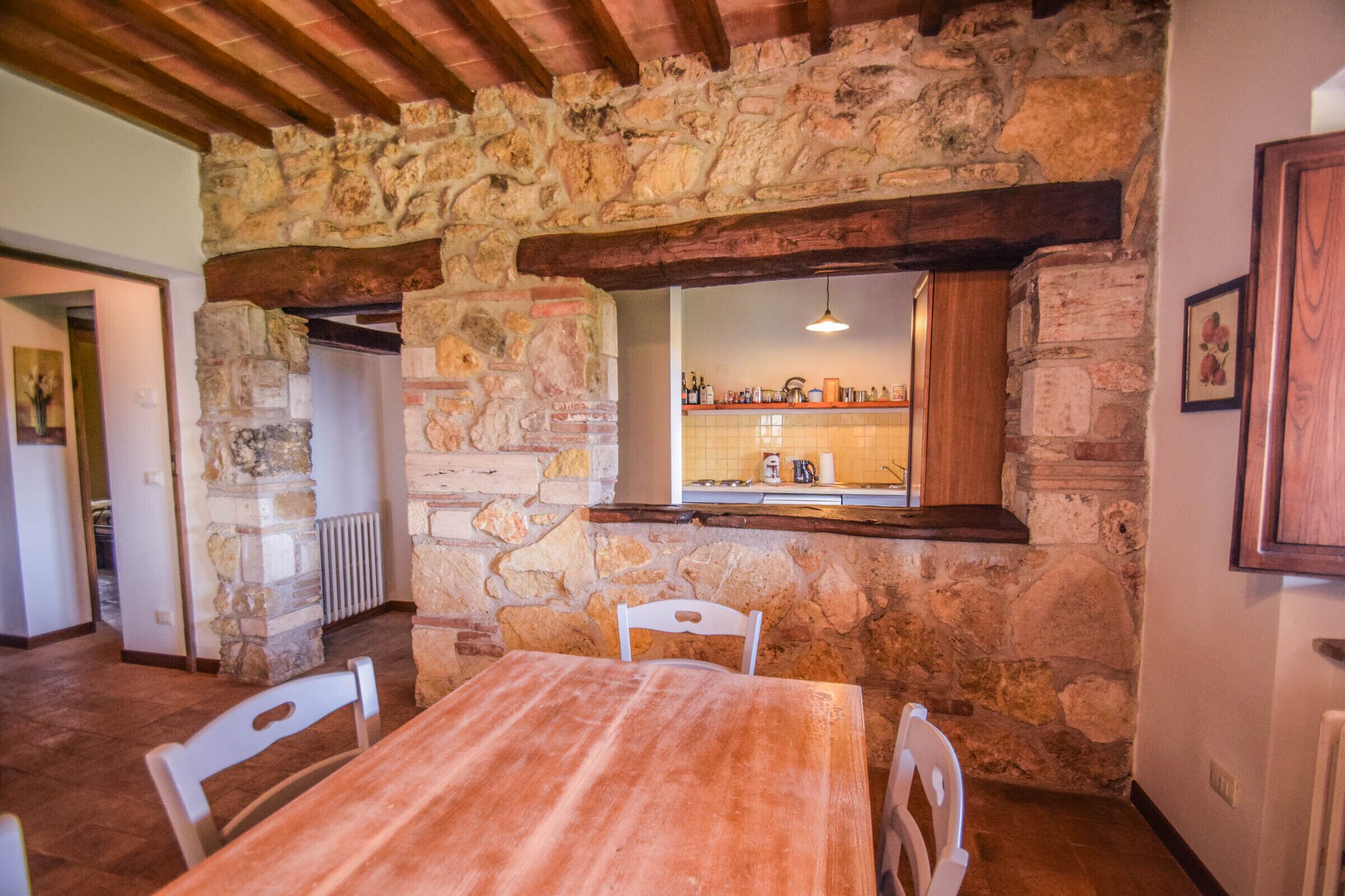Une pièce rustique aux murs de pierre, avec une table et des chaises en bois, une cheminée en pierre et un mur recouvert de carreaux dans le coin cuisine, visible à travers une fenêtre.