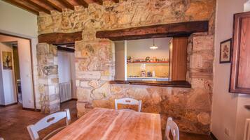 Une pièce rustique aux murs de pierre, avec une table et des chaises en bois, une cheminée en pierre et un mur recouvert de carreaux dans le coin cuisine, visible à travers une fenêtre.