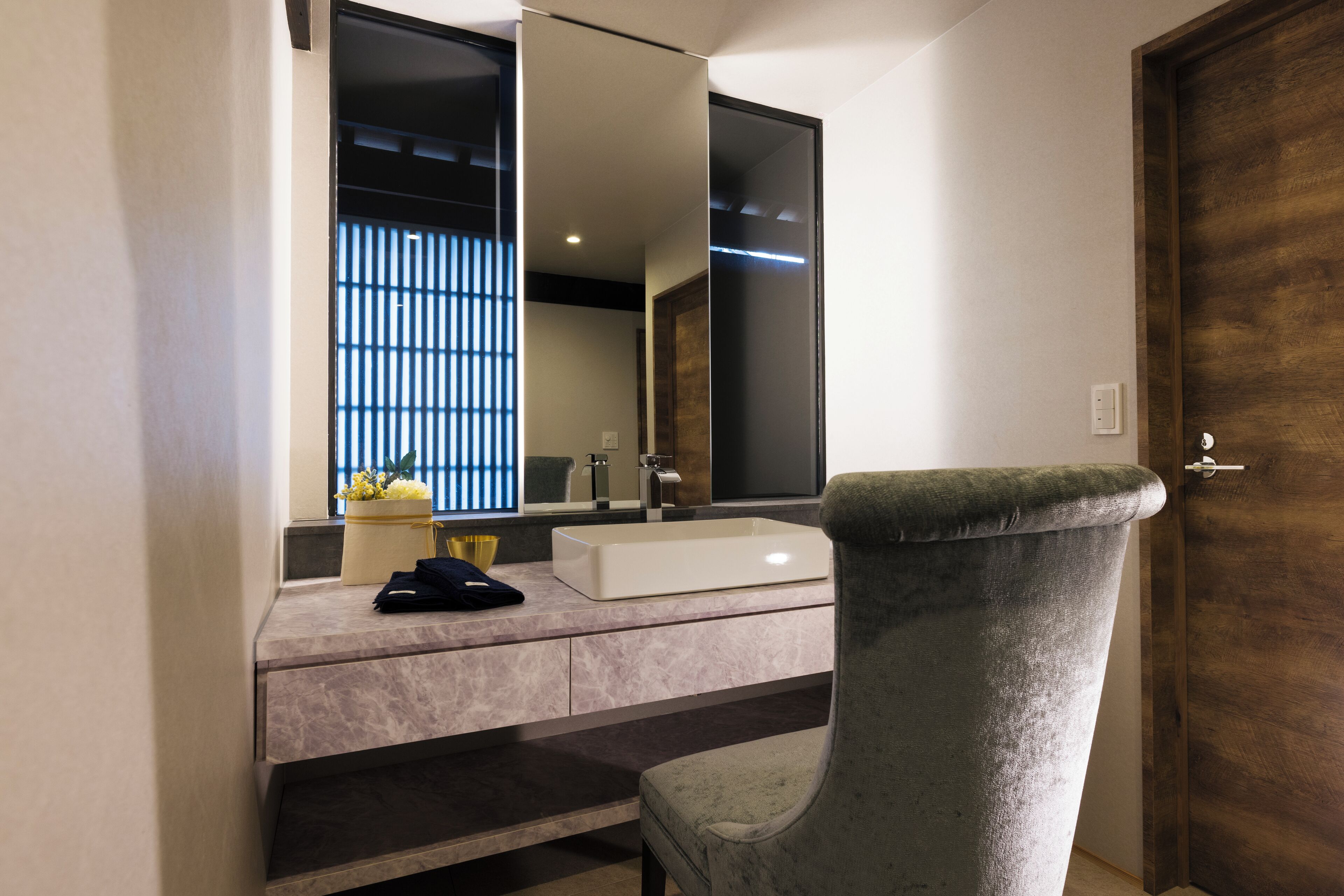 A modern bathroom with a large mirror, a white sink, and a grey chair.