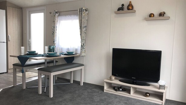 Una sala moderna con un televisor de pantalla plana sobre un soporte de madera, un comedor con mesa y sillas, y una cocina visible al fondo.