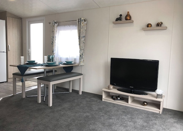 Un salon moderne avec un téléviseur à écran plat posé sur un meuble en bois, un coin repas avec une table et des chaises, et une cuisine visible au fond.