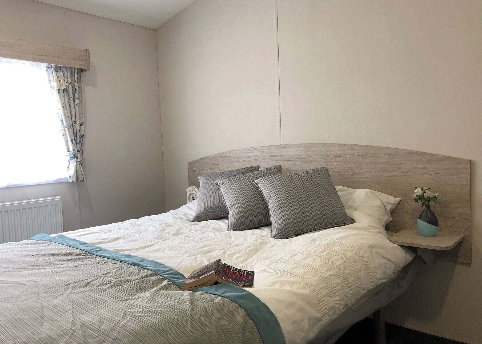 Ein ordentlich gemachtes Bett mit Kissen, ein Nachttisch mit einer Vase und ein Fenster mit Vorhängen.
