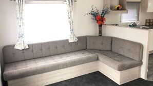 Un sofá en esquina con una ventana, un florero con flores y una zona de cocina.