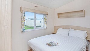 A bedroom with a bed, a window with curtains, a bedside table, and a wall-mounted shelf. - Caravan with one double and two twin. (Prestatyn, Denbighshire)