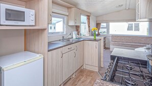 A compact kitchen with a microwave, sink, and stove. There are wooden cabinets and a window providing natural light. - Caravan with one double and two twin. (Prestatyn, Denbighshire)