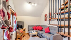 Un saló modern amb un sofà gris, coixinets vermells, un televisor de pantalla plana, una taula auxiliar de fusta i una escala amb baranes metàl·liques.