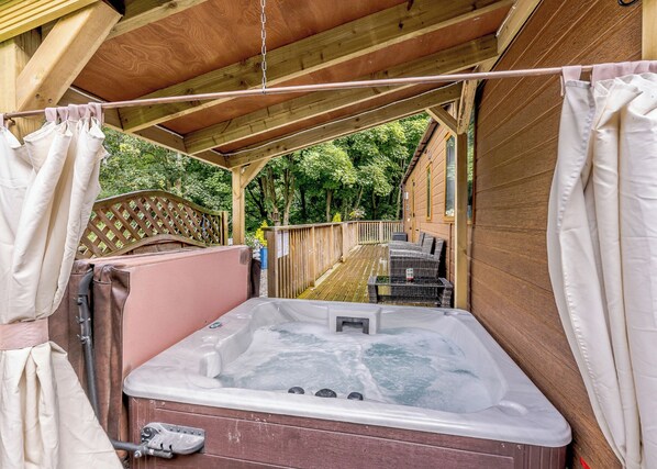 Un bain à remous installé sur une terrasse en bois à l’extérieur, avec une clôture en bois et de la verdure en arrière-plan.