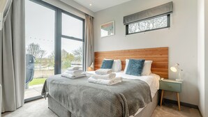 A modern hotel room with a large bed, a wooden headboard, a nightstand with a lamp, and a view of the outdoors.