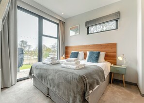 Una habitación de hotel moderna con una cama grande, un cabecero de madera, una mesita de noche con lámpara y vistas al exterior.