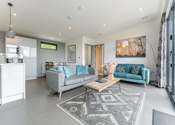 A modern living room with a grey sofa, a blue sofa, a coffee table, and a kitchen area with a microwave and a dining table.