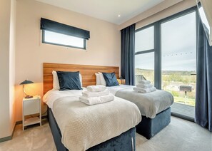Ein Hotelzimmer mit zwei Betten, einem Nachttisch, einer Lampe, einem Fenster mit Vorhängen und Blick ins Freie.