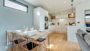 A modern kitchen with white cabinets, a dining area with a wooden table and copper chairs, and a grey sofa in a living area.