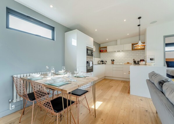 Une cuisine moderne avec des meubles blancs, un coin repas doté d’une table en bois et de chaises en cuivre, et un canapé gris dans l’espace salon.