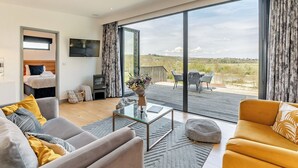 A modern living room with a sofa, armchair, and a coffee table. There is a TV mounted on the wall, and a balcony with outdoor seating.
