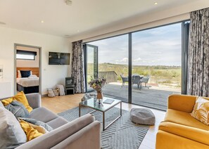 Un salon moderne avec un canapé, un fauteuil et une table basse. Un téléviseur est fixé au mur, et il y a un balcon avec des sièges d’extérieur.