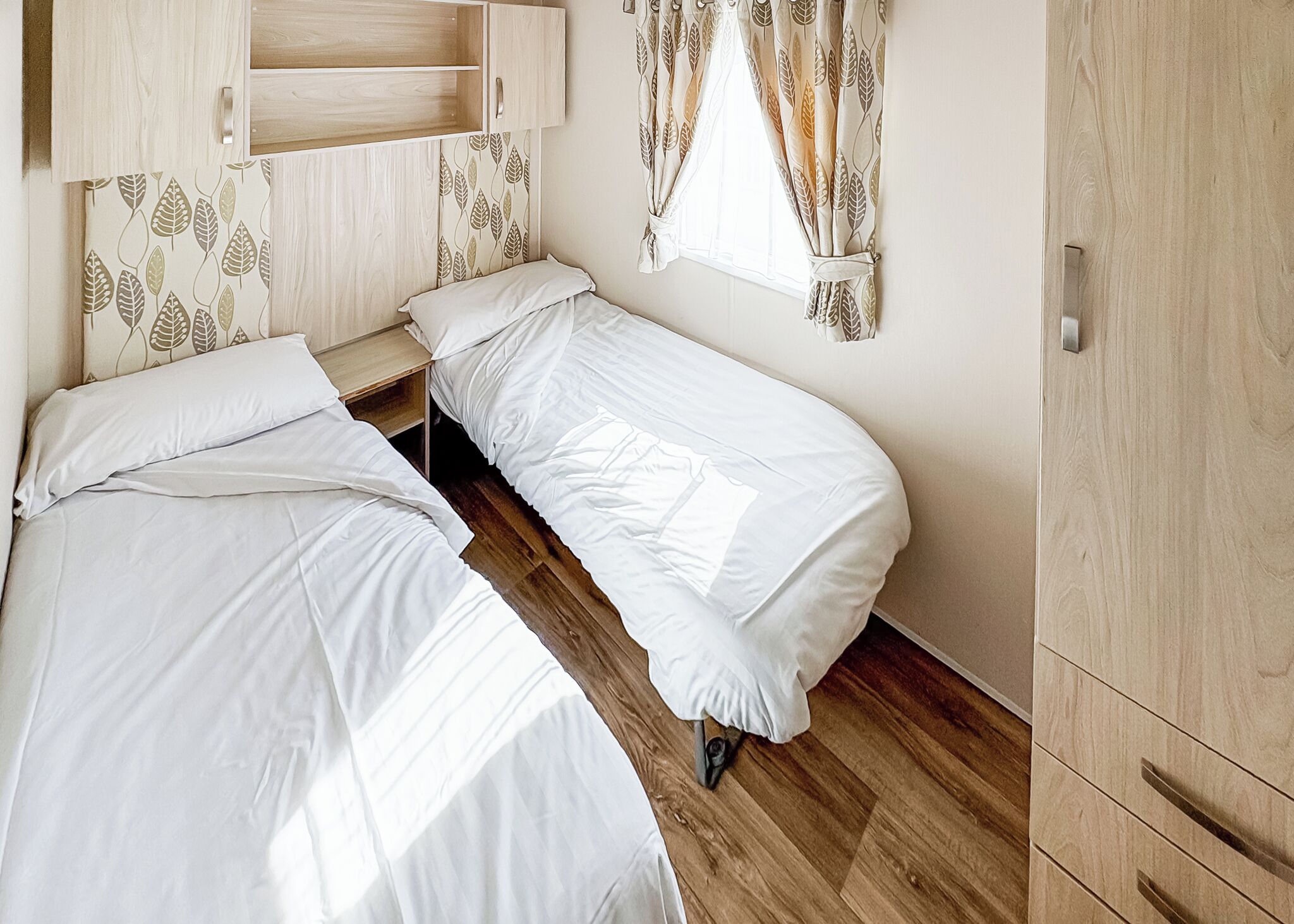 Una habitación con dos camas individuales, un armario de madera y una ventana con cortinas de hojas.