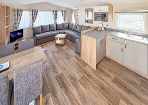 A modern RV interior with a sectional sofa, dining table, kitchen area, and a TV.