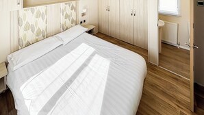 A bedroom with a bed, wooden wardrobe, bedside table, and a window. - Modern and contemporary 12ft-wide caravan with one double and one twin. (Bleadon, Weston-Super-Mare)