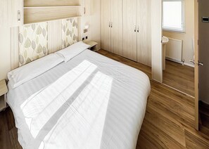 A bedroom with a bed, wooden wardrobe, bedside table, and a window. - Modern and contemporary 12ft-wide caravan with one double and one twin. (Bleadon, Weston-Super-Mare)