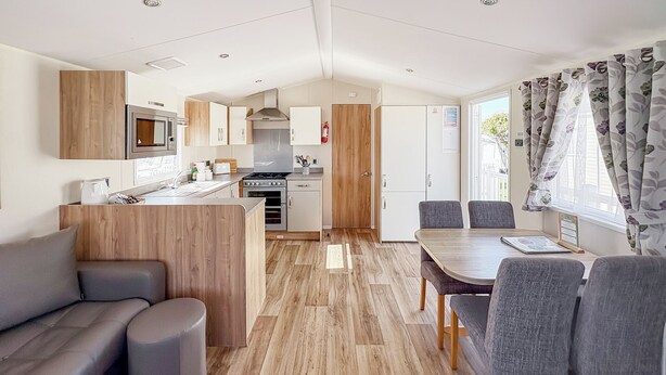 A modern kitchen with a microwave, stove, and oven, a dining area with a table and chairs, and a living area with a sofa and ottoman. - Modern and contemporary 12ft-wide caravan with one double and one twin. (Bleadon, Weston-Super-Mare)