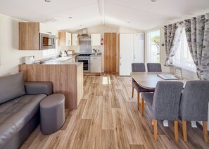 A modern kitchen with a microwave, stove, and oven, a dining area with a table and chairs, and a living area with a sofa and ottoman. - Modern and contemporary 12ft-wide caravan with one double and one twin. (Bleadon, Weston-Super-Mare)