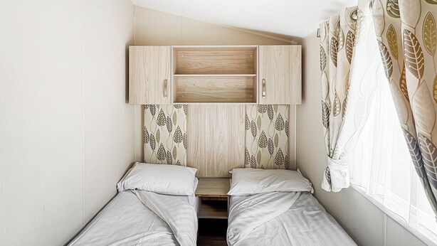 A bedroom with two single beds, a wooden wardrobe, leaf-patterned curtains, and a window with sheer white curtains.