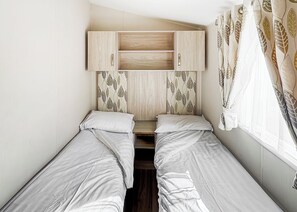 A bedroom with two single beds, a wooden wardrobe, leaf-patterned curtains, and a window with sheer white curtains. - Modern and contemporary 12ft-wide caravan with one double and one twin. (Bleadon, Weston-Super-Mare)