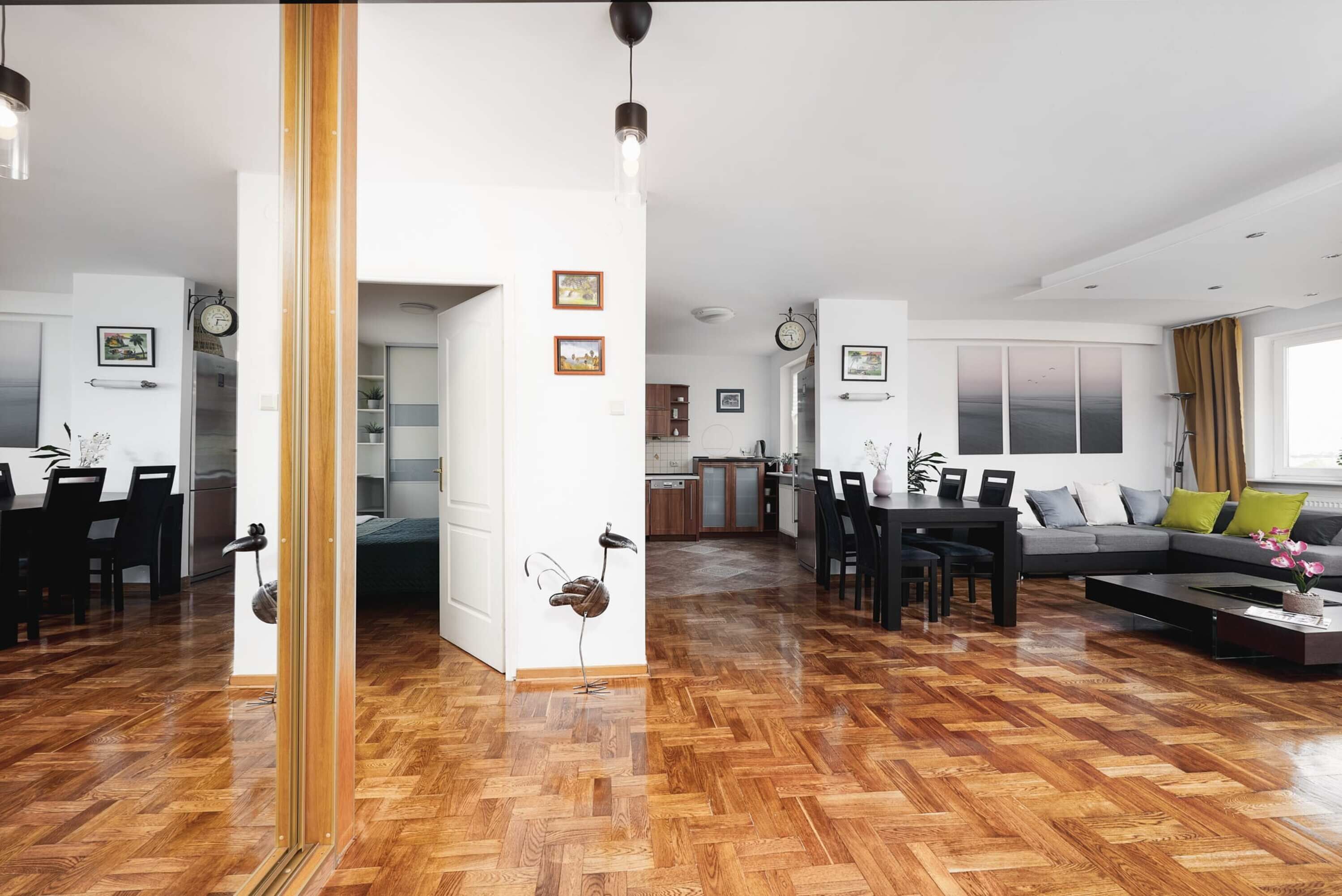 Uma sala de estar espaçosa com piso de parquet de madeira, uma mesa de jantar, um sofá e uma cozinha ao fundo.