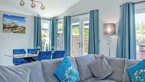 A modern living room with a grey sectional sofa, blue curtains, a dining table with chairs, and a painting on the wall. - Chatsworth Superior Pet Friendly (Longnor, Nr. Buxton)