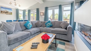 A modern living room with a grey sectional sofa, a wooden coffee table with remote controls and a vase of red roses, and a fireplace. - Chatsworth Superior Pet Friendly (Longnor, Nr. Buxton)