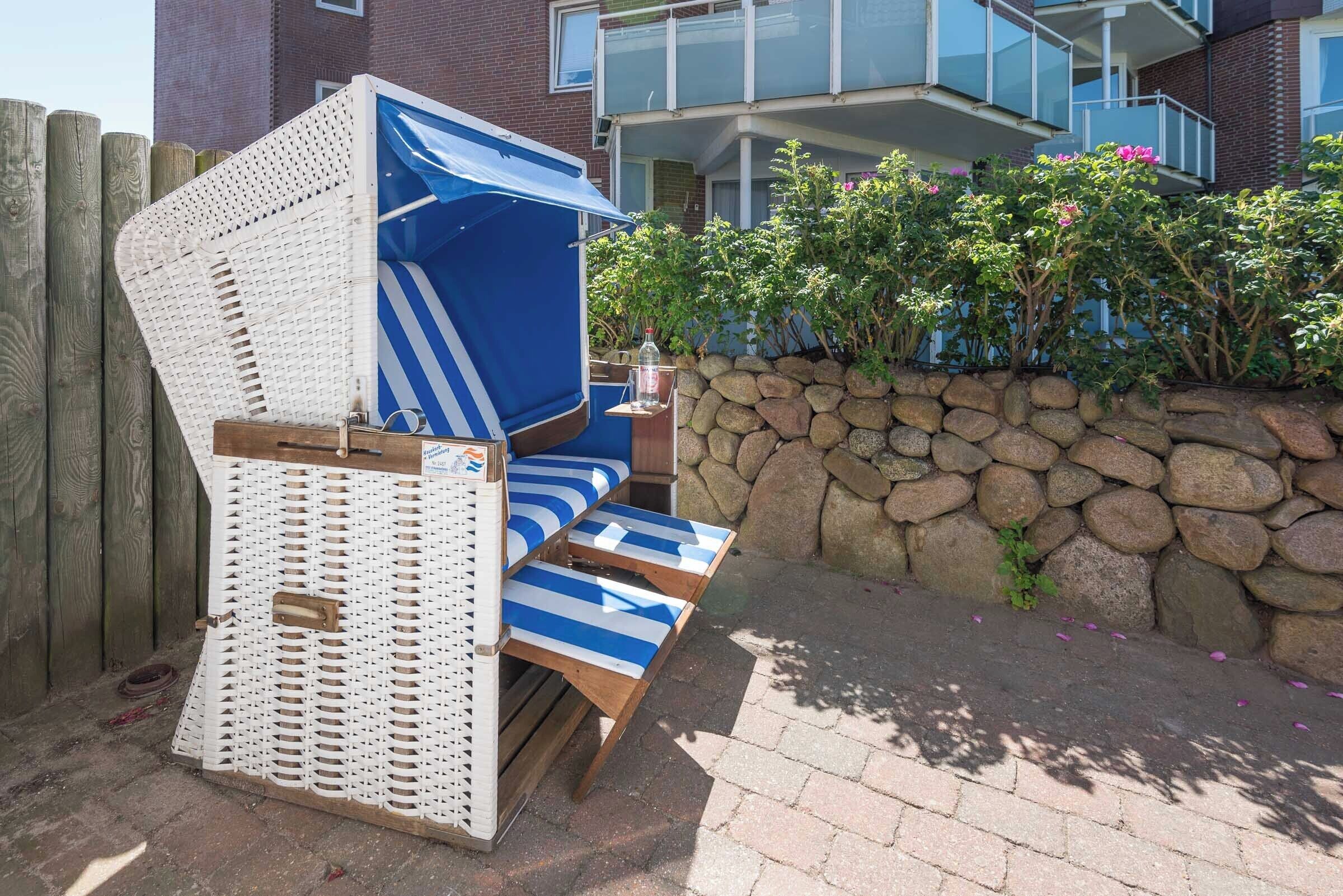 Un espace de détente extérieur aménagé avec des meubles en osier, des coussins à rayures bleues et blanches, un mur en pierre et une clôture en bois.