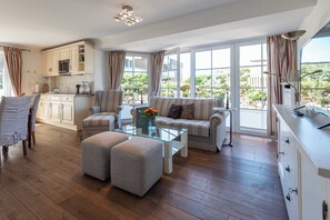 Ein geräumiges Wohnzimmer mit einer Couch, Hocker und einem Glastisch. Im Hintergrund ist eine Küche mit einer Mikrowelle und Schränken zu sehen.